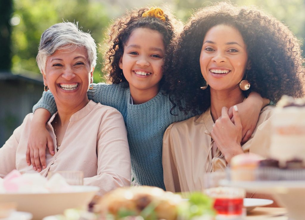 Three generations smiling and hugging outdoors at a family birthday party, with desserts on the table in the foreground.