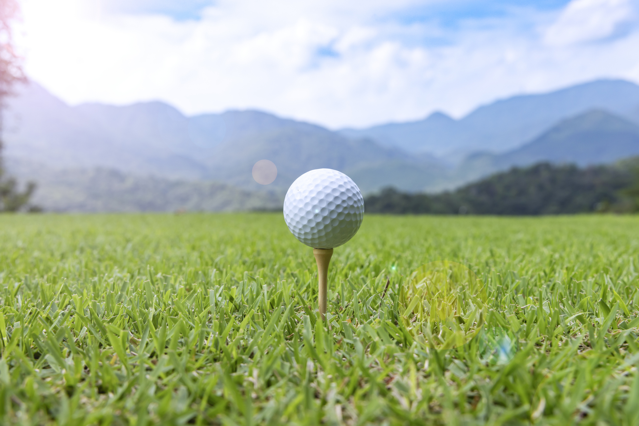 Golf ball on a tee in the foreground with a blurred mountain landscape and blue sky behind.