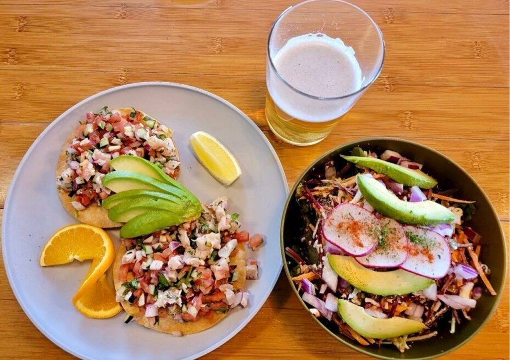 CHIP_Eatery66 - Chipeta Lodge Resort + Spa Tacos with pico de gallo and avocado slices on a plate, lime and orange wedges nearby, plus a beer and a veggie salad on a wooden table.
