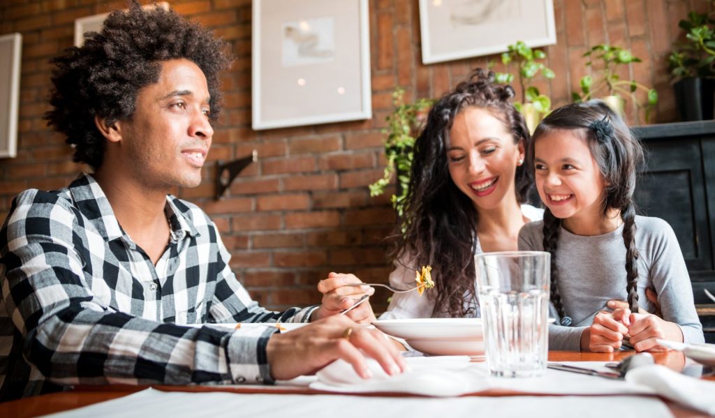 CHIP_Family-Dining - Chipeta Lodge Resort + Spa A man in a plaid shirt sharing a meal with a smiling woman and a young girl at a brick-walled cafe table.