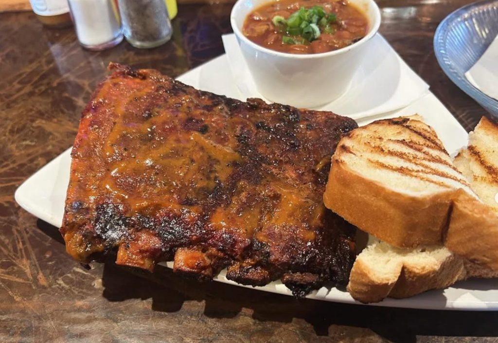 CHIP_Lazy-Dog-Saloon - Chipeta Lodge Resort + Spa Rack of glazed barbecue ribs on a white plate with toasted bread and a bowl of chili in the background, on a wooden table.