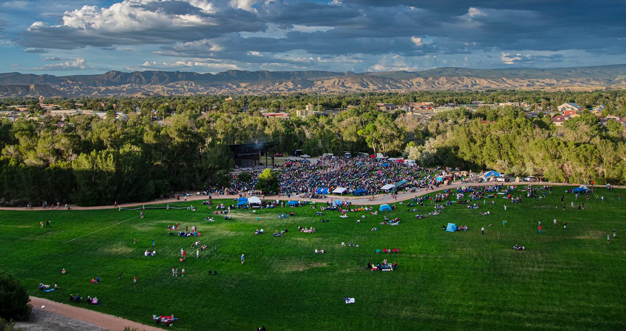 Aerial view of a crowded outdoor festival in a green park with a large stage, tents, and people spread across the grass, mountains in the distance under a cloudy sky.