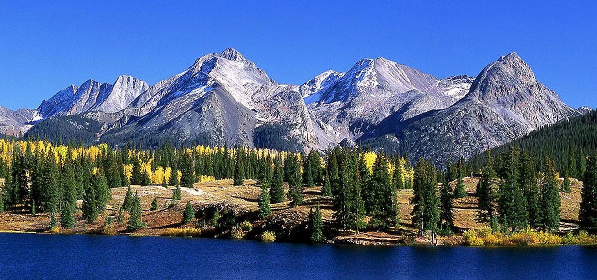 Chipeta_San-Juan-Skyway - Chipeta Lodge Resort + Spa Snow-capped mountains beyond a forest of evergreens with a calm blue lake in the foreground under a clear sky, fall colors visible on the trees.