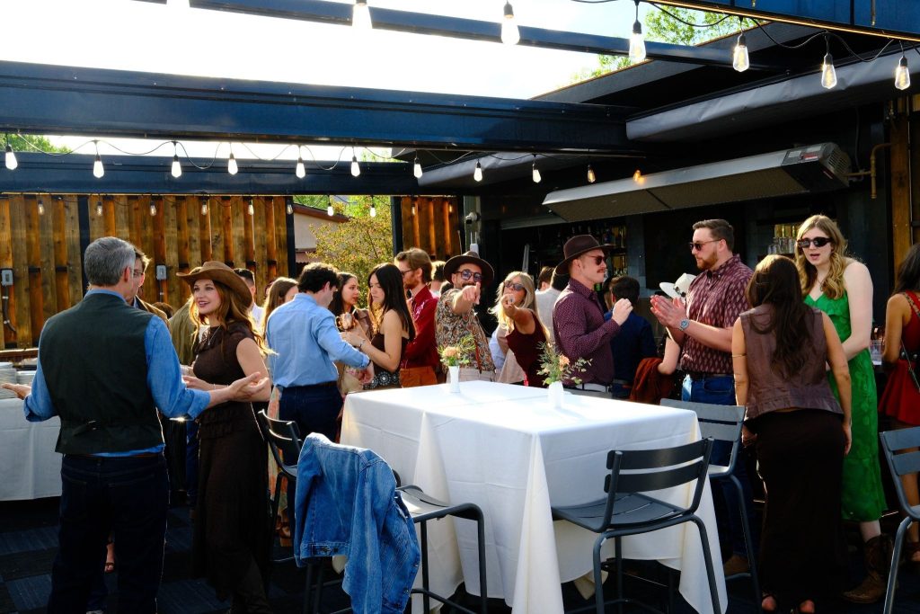 A lively outdoor party with groups of people mingling under string lights above, standing around tables with white tablecloths in a patio setting.