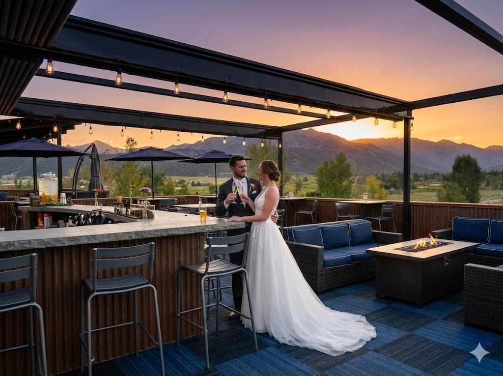 Bride and groom toast at an outdoor bar on a sunset terrace, with mountains in the distance.