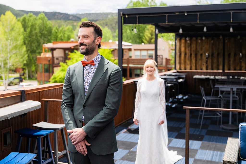 Man in a gray suit with an orange bow tie and floral shirt stands on a wooden deck; a bride in a white wedding dress follows behind on an outdoor venue.