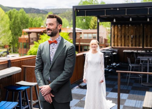 Man in a gray suit with an orange bow tie and floral shirt stands on a wooden deck; a bride in a white wedding dress follows behind on an outdoor venue.