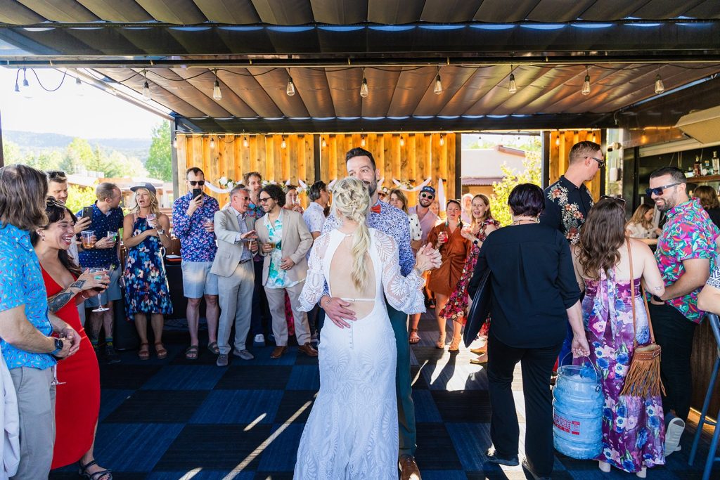Bride and groom dance center stage as guests toast and chat at an outdoor wedding reception under a covered patio.