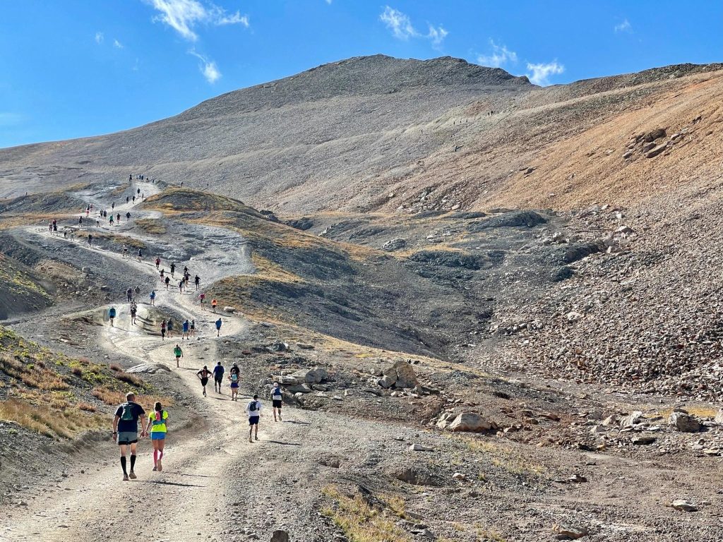 image - Chipeta Lodge Resort + Spa Group of hikers walking along a rocky mountain trail up a barren slope under a clear blue sky.