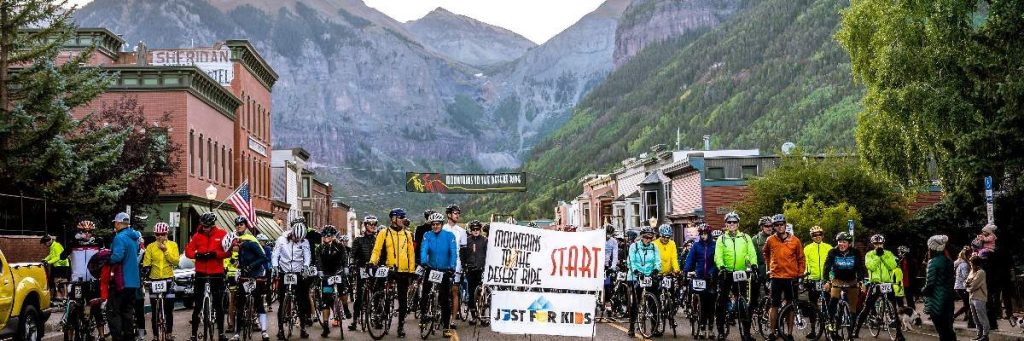 image - Chipeta Lodge Resort + Spa Group of cyclists in bright jackets line up at a mountain-town street start, with banners and mountains in the background, ready to ride.